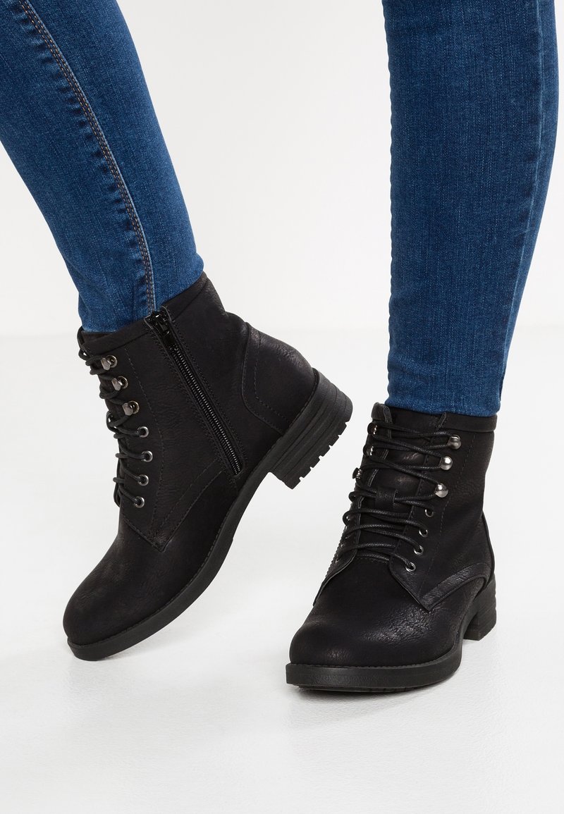 Personne portant des bottines noires à lacets avec des fermetures éclair sur le côté, associées à un jean skinny bleu sur fond blanc.