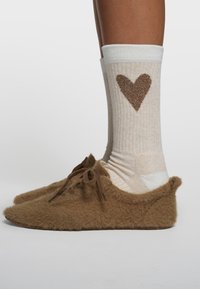 Beine mit cremefarbenen Socken mit braunem Herz, kombiniert mit braunen, flauschigen Hausschuhen mit Schnürsenkeln, vor weißem Hintergrund gezeigt.