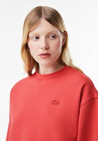 Röd sweatshirt gjord av mjukt material, med rund halsringning och en liten, prägling logotyp p å vänster bröst. Slät textur med långärmar.