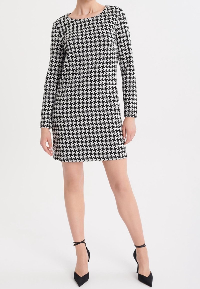 Robe à motif pied-de-poule noir et blanc, manches longues, col rond, longueur au genou, en tissu texturé, associée à des chaussures à talons noirs.