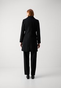 Abrigo negro de lana con cuello muesca, corte ajustado y espalda con ventilación, combinado con pantalones negros y calzado de tacón, mostrado desde la parte trasera.