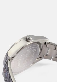 Edelstahluhr mit runder Gehäuseform, strukturiertem Lünettenrand und einem gemusterten Armband. Die Rückseite zeigt gravierte Designs und eine auffällige Krone.