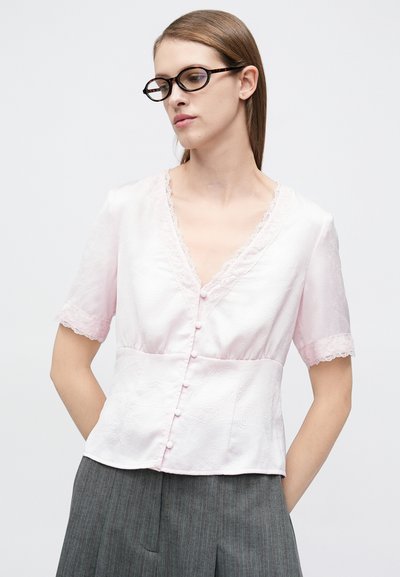 Ροζ σατέν μπλούζα με κοντά μανίκια, δαντέλα στο λαιμό και στα μανίκια, και κουμπιά στο μπροστινό μέρος. Φορεμένη με γκρι πλισέ φούστα.