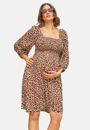 Gravid kvinde iført brun og beige leopardprint kjole og mørke solbriller, hviler den ene hånd på maven og den anden på taljen.
