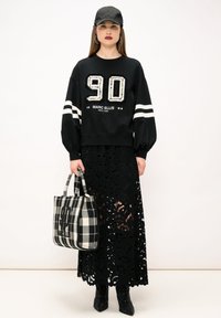 Sweatshirt noir avec "90" en strass et rayures blanches, associé à une jupe noire en dentelle. Tient un sac fourre-tout à carreaux en noir et blanc.