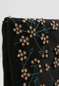 Pochette en tissu noir ornée d'une broderie florale complexe en fils dorés et argentés, avec des accents floraux en relief et des coutures vertes.