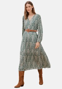 Maxikleid mit grünem Paisley-Druck, langen Ärmeln, V-Ausschnitt und einem braunen Gürtel in der Taille. Getragen mit braunen Stiefeln bis zum Knie.