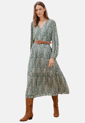 Maxikleid mit grünem Paisley-Druck, langen Ärmeln, V-Ausschnitt und einem braunen Gürtel in der Taille. Getragen mit braunen Stiefeln bis zum Knie.