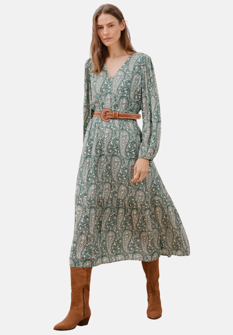 Maxikleid mit grünem Paisley-Druck, langen Ärmeln, V-Ausschnitt und einem braunen Gürtel in der Taille. Getragen mit braunen Stiefeln bis zum Knie.