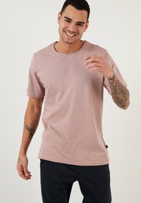 Tricou scurt cu mânecă scurtă de culoare roz deschis, cu guler rotund, textură moale și fante laterale. Prezintă tatuaje vizibile pe brațele modelului.