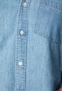 Ljusblå denimskjorta med spetsig krage, framficka och vita knappar. Tyget har en subtil struktur och en urblekt look.