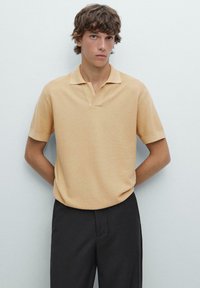 Polo shirt beige a maniche corte realizzato in un tessuto testurizzato, con colletto classico e vestibilità rilassata, abbinato a pantaloni neri.