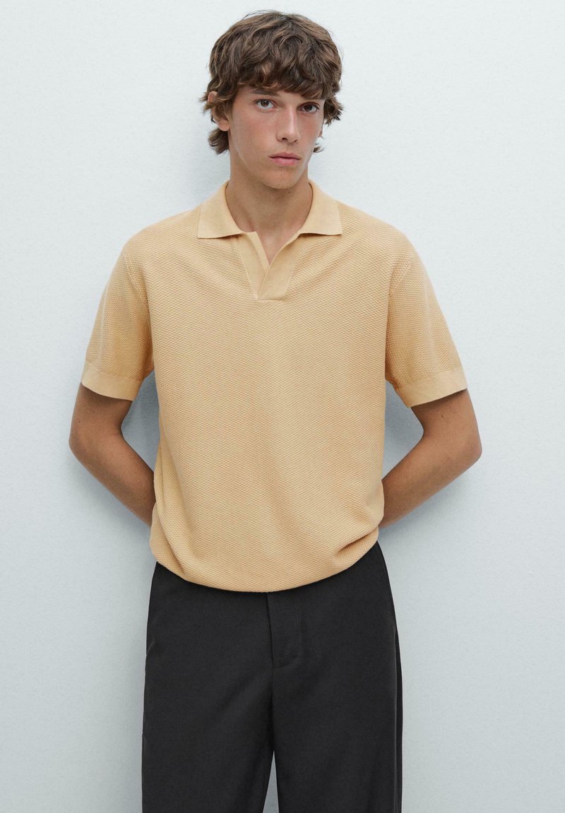 Polo shirt beige a maniche corte realizzato in un tessuto testurizzato, con colletto classico e vestibilità rilassata, abbinato a pantaloni neri.