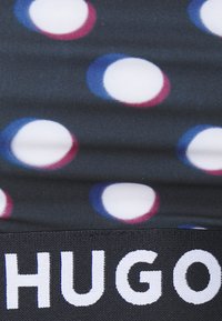 Crni materijal s višebojnim kružnim uzorcima u bijeloj, plavoj i ružičastoj boji, s istaknutim "HUGO" logotipom duž donjeg dijela.