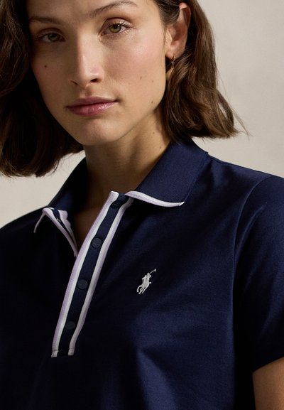 Γυναίκα με κοντά καστανά μαλλιά που φοράει μπλε σκούρο πόλο μπλούζα με λευκή ρίγα στο γιακά και μικρό κεντημένο λογότυπο με ιππέα αλόγου.