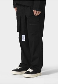Sorte cargo bukser har en løs pasform, flere lommer, et label patch foran, og de er parret med sorte sneakers med hvide snørebånd.