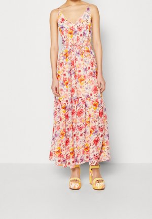 Kobieta w różowej, kwiecistej, maxi sukience na ramiączkach z dekoltem w serek, z wiązanym paskiem w talii oraz w żółtych, kwiecistych sandałach na wysokim obcasie z platformą.