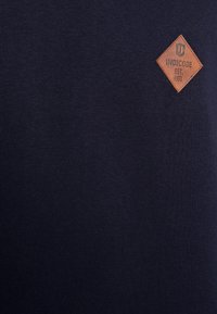 Patch marron en forme de diamant avec "INDICODE EST. 1993" sur fond en tissu texturé bleu foncé.