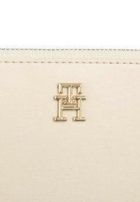 Cremfarbener Lederwallet mit einem Logoemblem in Goldton. Die Oberfläche hat eine glatte Textur und feine Nähte entlang der Kanten.