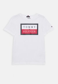 White cotton t-shirt featuring a rectangular graphic with "TOMMY HILFIGER" text in black, red, and white colors. Short sleeves, crew neck design.