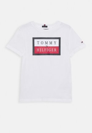 Bílý bavlněný tričko s obdélníkovou grafikou a textem "TOMMY HILFIGER" v černé, červené a bílé barvě. Krátké rukávy, kulatý výstřih.