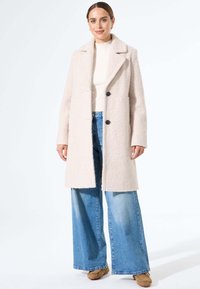 Beige textuurjas met een geknikte kraag, twee zwarte knopen, gecombineerd met brede blauwe denim jeans en beige schoeisel.