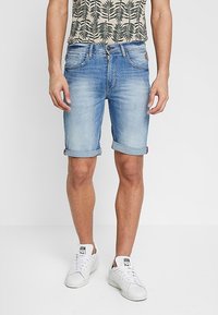 Ljusblå denimshorts med upprullade fållar, utrustade med fem fickor och en kontrasterande logga. Bärs tillsammans med en mönstrad kortärmad skjorta.