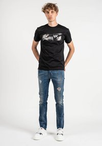 Camiseta de algodón negra con texto gráfico metálico "LES HOMM" en el frente, combinada con jeans azules desgastados y zapatillas blancas. Corte casual.