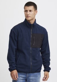Marineblauwe fleece jas met een zwarte ritsborstzak, hoge kraag en zijzakken. Beschikt over een gladde textuur en contrasterende materialen.
