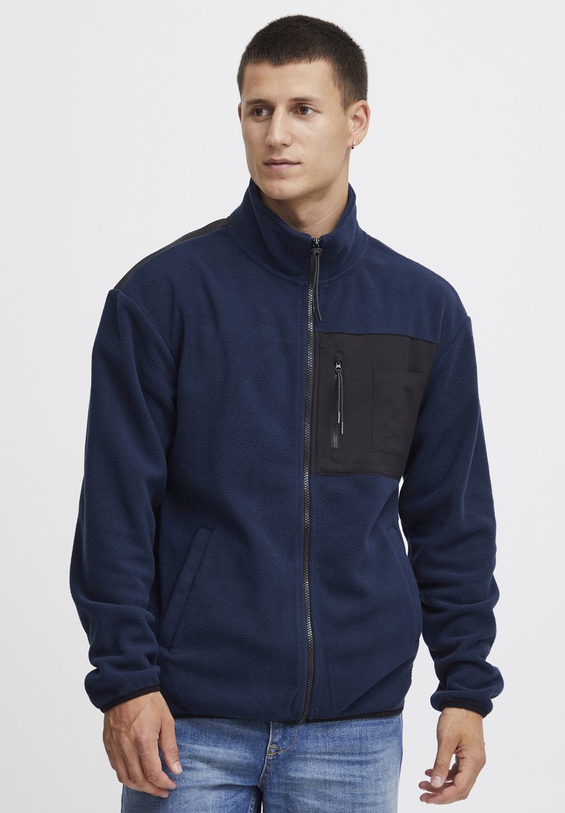 Marineblauwe fleece jas met een zwarte ritsborstzak, hoge kraag en zijzakken. Beschikt over een gladde textuur en contrasterende materialen.