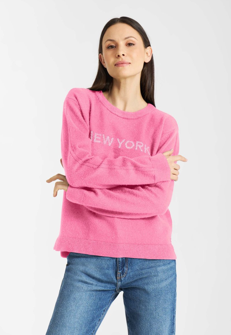 Różowy sweter z polaru z napisem "NEW YORK" w białym kolorze na klatce piersiowej, okrągłym dekoltem i ribbowanym dołem. Połączony z niebieskimi dżinsami.