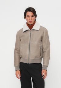 Beige műbőr bomber dzseki fehér gyapjú gallérral, elöl cipzárral, oldalsó zsebekkel és bordás mandzsettákkal. Barna garbó fölé hordva.
