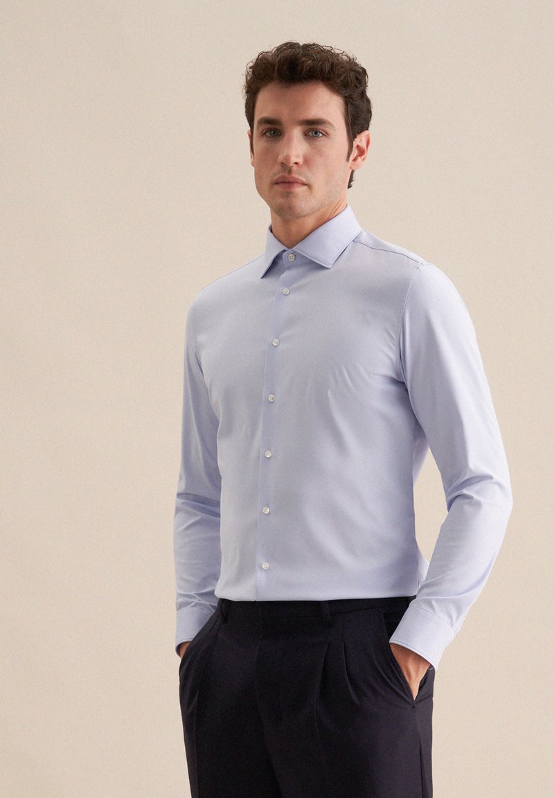 Homme portant une chemise bleu clair à manches longues et un pantalon noir, debout les mains dans les poches devant un fond beige uni.