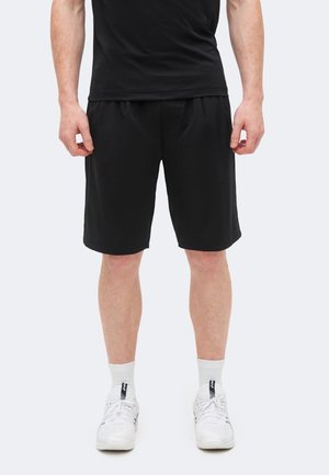 Άνδρας που φοράει μαύρο αθλητικό σορτς, μαύρο μπλουζάκι, άσπρες κάλτσες και άσπρα αθλητικά παπούτσια, στέκεται μπροστά σε λευκό φόντο.