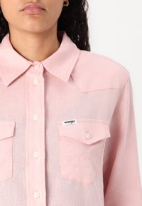 Camisa botão rosa claro com colarinho, dois bolsos no peito e logótipo Wrangler. Apresenta um tecido texturizado subtil e botões semelhantes a pérolas.