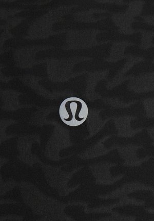 Svart tyg med ett texturerat kamouflagemönster; har en cirkulär silverlogotyp centrerad på materialet.