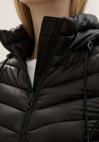 Chaqueta acolchada negra con cuello alto, que cuenta con cierre mediante cremallera, textura acanalada y detalles con cordón alrededor de la capucha.