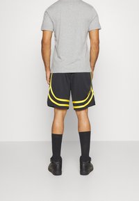 Shorts de sport noirs avec des bandes courbes jaunes, associés à un t-shirt gris, des chaussettes noires hautes jusqu'aux genoux et des chaussures noires. Vue arrière.