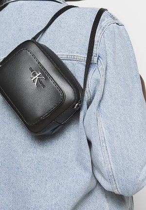 Sac bandoulière en cuir noir avec fermeture éclair, logo argenté, et sangle réglable, porté avec une veste en jean claire.