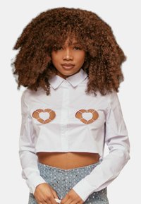 Camicia bianca corta con colletto, caratterizzata da tagli a forma di cuore in un design marrone sul busto. Tessuto di cotone liscio.