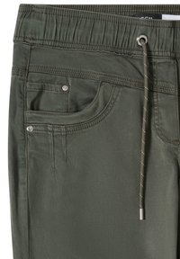 Gros plan sur un pantalon vert olive avec une taille élastique, un cordon de serrage et une poche avant ornée de rivets métalliques et de détails de couture.