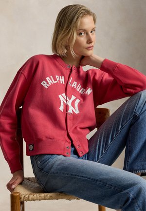 Jonge vrouw in een rode Ralph Lauren sweater met knopen en blauwe spijkerbroek, zittend op een houten stoel met een gevlochten zitvlak, leunt met haar hoofd op haar hand.