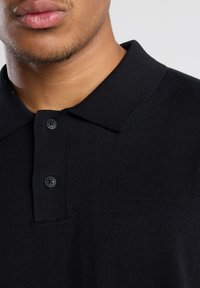 Black short-sleeve polo shirt with a textured knit fabric, buttoned placket, and a classic collar. Accents include two black buttons.
