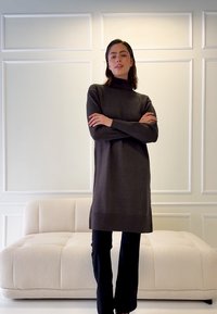 Gestricktes dunkelgraues Rollkragenkleid mit lockerem Sitz, geripptem Saum, kombiniert mit schwarzer Hose. Schlichtes Design vor hellem, getäfeltem Hintergrund.