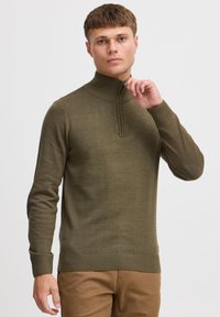 Pull vert olive avec col haut et fermeture éclair. Dot�é de poignets et d'un ourlet côtelés, avec des coudières. Porté avec un pantalon khaki.