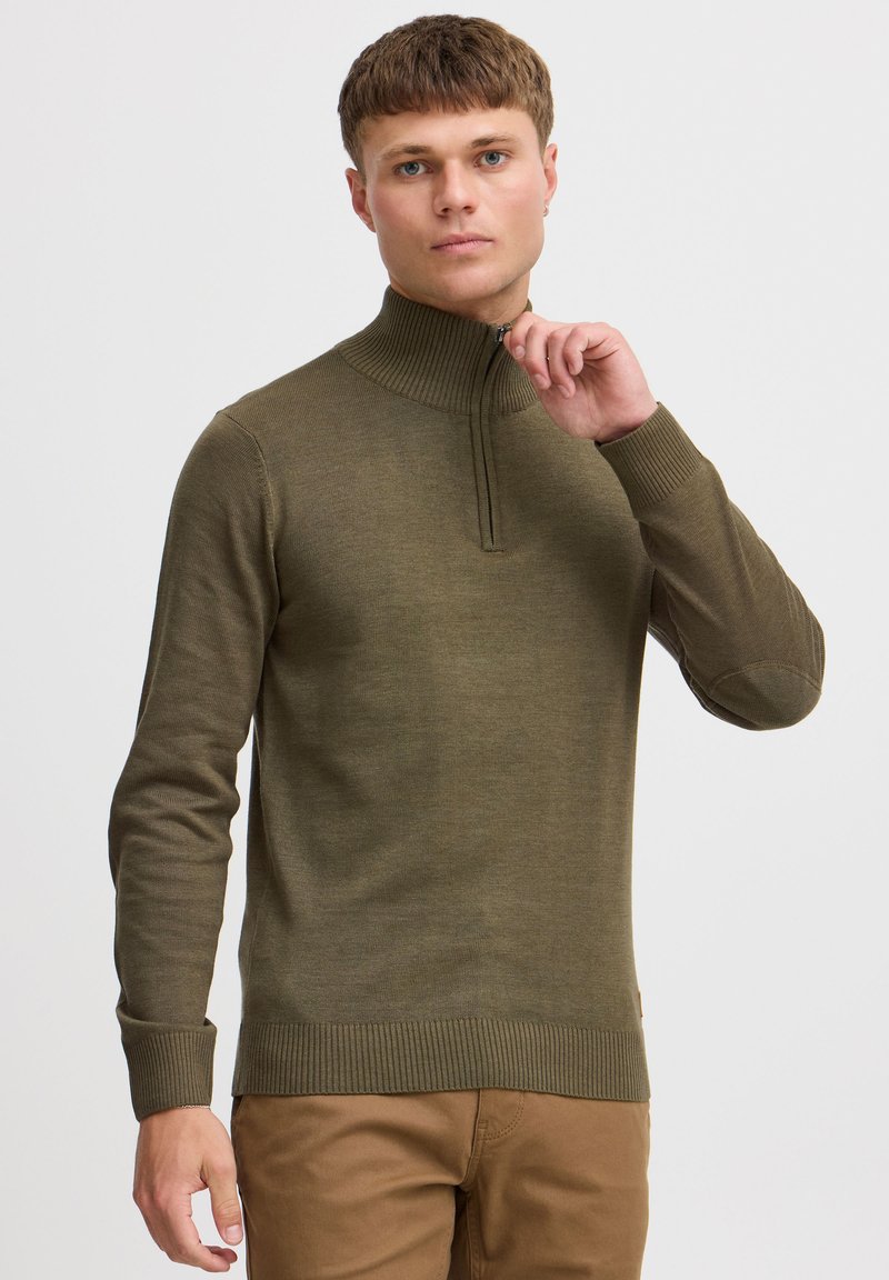 Pull vert olive avec col haut et fermeture éclair. Doté de poignets et d'un ourlet côtelés, avec des coudières. Porté avec un pantalon khaki.