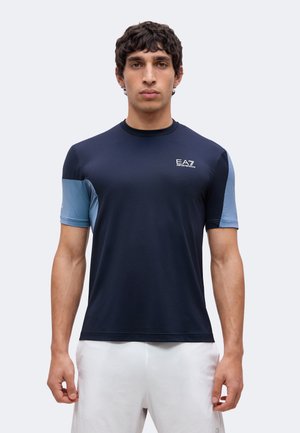 Tânăr privit din față purtând un tricou sportiv cu mânecă scurtă bleumarin și albastru deschis cu logo EA7 și pantaloni scurți albi.