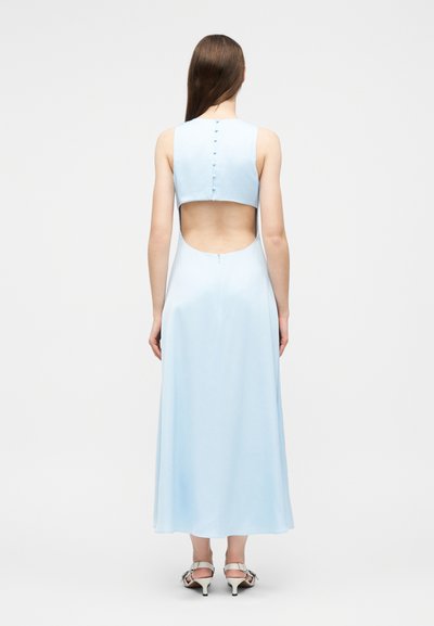 Femme aux longs cheveux bruns portant une robe bleu clair sans manches avec un dos boutonné et ouvert au milieu du dos, assortie à des talons blancs à brides.