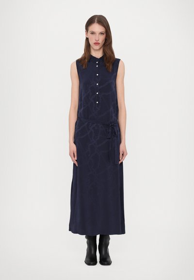 Ärmelloses, marineblaues Kleid mit Kragen, Knopfleiste und gebundenem Taillenbereich, das eine subtile gemusterte Textur aufweist. Getragen mit schwarzen Ankle-Boots.