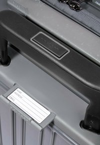 Valise rigide grise avec une surface texturée. Elle est équipée d'une poignée en plastique noir et d'un porte-étiquette sur le devant, avec un espace pour les coordonnées.